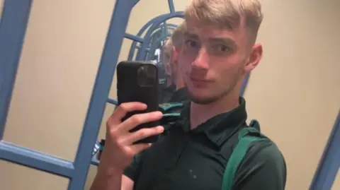 Facebook Jamie Kadolski takes a selfie in a mirror. He has short blond hair and is wearing a green polo neck with green epaulettes on either shoulder. His phone is black.  