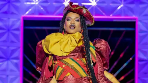 BBC/World of Wonder/Guy Levy Rileasa Slaves on the runway. She wears a red pirate-style hat, long braids and a red puff-sleeved dress matched with yellow scarf and a cropped waist coat in red, yellow, green and gold stripes.