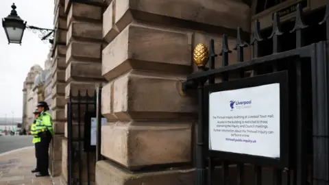 EPA A Liverpool City Council sign with details about the inquiry's access and restrictions plus weblink for extra information is seen on a gate at the town hall, with two police officers in yellow hi-vis jackets standing guard outside the Georgian building