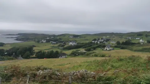 A general view of Sleat. There is a barbed wire fence and stone dyke in the foreground. There are houses dotted across the landscape and the sea in the distance.