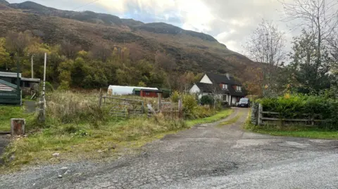 A house at the end of a track. There is a van parked outside it. Behind the house is a hill.