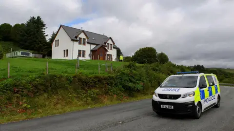 PA Media A police van parked below a white-walled detached property. A police officer is near the house.