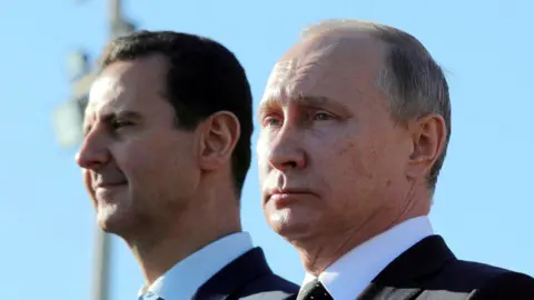 Reuters Putin and Assad stand next to each other. Both are wearing suits, with the sky acting as background