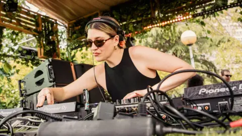 Getty Images Moxie during a set. Moxie has dyed blonde hair which is tied back. She wears a black vest top and sunglasses, headphones over one ear as she leans over her deck. She's performing outside on a sunny day, with green foliage behind her. 