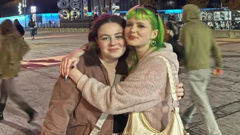 Freya and Niamh outside the Co-op Live Arena in Manchester. Niamh has dyed green hair and has her arms around Freya, who nods to the Brat theme with green eye shadow. They're both wearing brown coats and smile at the camera. 