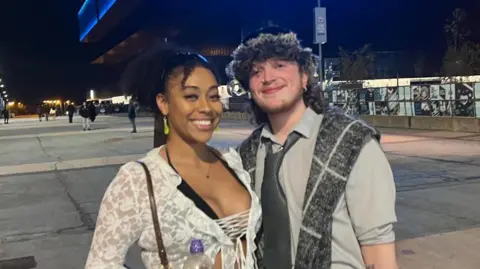 Tasha and Lucas outside the Co-op Live Arena in Manchester. Tasha wears a Brat inspired outfit - a sheer white top tied together at the front. Lucas wears a grey shirt with a thick dark grey tie and a grey tartan scarf over his shoulders. 