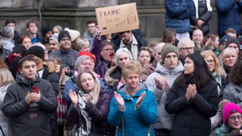 PA Media Dozens of mourners applauding as Janey Godley's hearse passes by. One of them is holding up a sign on cardboard saying 'Frank! Get the gates' with a drawing of gates.