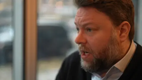 A man with a thick beard is talking to an interviewer who is off-camera. The man has dark brown hair and is wearing a smart white shirt with a dark jumper over the top of it. He is sat next to a window, through which the pavement and a passing vehicle is visible.