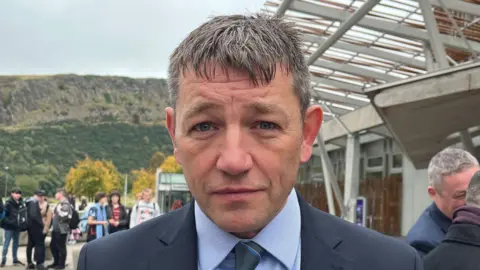 David Threadgold standing outside the Scottish Parliament building. He is looking straight into the camera. He is wearing a suit, with a light blue shirt and dark tie. 