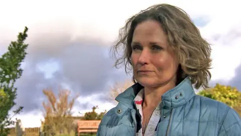 Rhiannon Bragg standing outside on an autumn day, with trees behind her. She is looking concerned and wearing a light blue or aqua jacket.