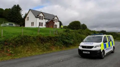 PA Media A police van on the road outside the home of the MacKinnons, where John MacKinnon was shot dead by his brother in law. The home has white walls, with a brown doorway and entrance, while there is greenery in a large garden outside it. 