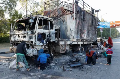 Reuters Women and children collect recyclables from the burnt truck used by Bushra Bibi, wife of jailed former Pakistani Prime Minister Imran Khan. The truck is in the middle of a quiet main road