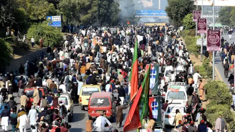 Reuters Hundreds of people make their way along a highway with bushes on either side, and handful of cars in amongst the protesters. Some people hold giant red and green flags. Smoke can be seen rising in the distance.