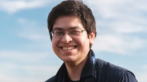 Husban Siddiqui A young man in glasses smiles widely against a cloudy blue sky. The sunlight hits the side of his face, suggesting a late autumn afternoon. 