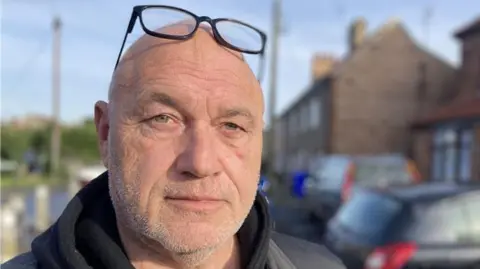 Matt Precey/BBC Johnny Whitfield smiles at the camera with black glasses that rest on his head. He is standing on a residential street which is blurred behind him.