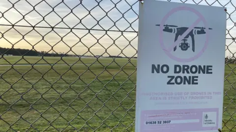 Matt Precey/BBC A close up of a sign on a silver fence that reads "No drone zone", with an image of a drone with a red circle cross. An airfield can be seen behind the fence.