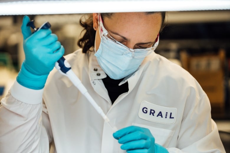 A man wearing a white lab coat with the word 'Grail' holds a pipette. 