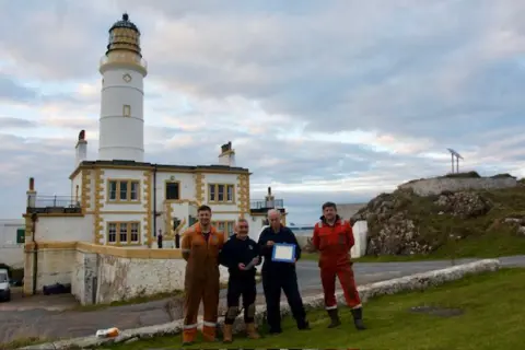 Ross Russell Ross, two engineers and the lighthouse keeper stand outside Corsewall Lighthouse. They are wearing workmen jumpsuits. The lighthouse is white.