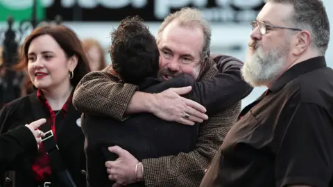 PA Media Gavin Mitchell hugs a man in the middle of shot, at the right is a large man with grey heair and glasses and  a younger woman with red lipstick on the left. All are wearing dark colours.