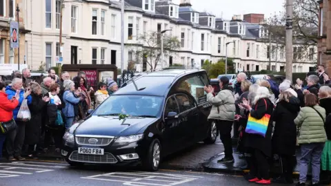 PA Media A black hearse in the middle of a road with  a small crowd of people wearing warm looking jackets and carrying bags clapping their hands.