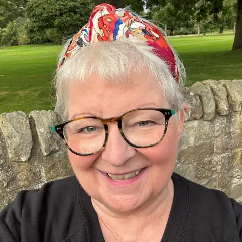 PA Media Janey Godley, with short grey hair, dark glasses and a red bandana on her head, smiles at the camera with a stone wall and green park behind her 