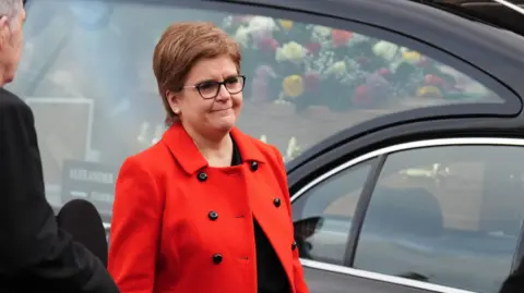 PA Media Sturgeon wearing a bright read coat stands in front of a hearse with a coffin covered in flowers.