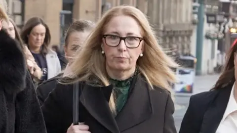 A woman with blonde hair and black glasses, wearing a black coat and green top, has a stern face as she looks at the camera while walking along a pavement with members of her legal team.