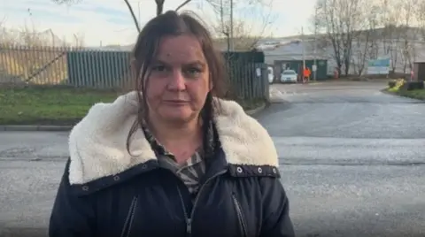 Rebecca Currie is standing in front of a road. She has brown hair tied in a plait, and is wearing a black coat with white fuzzy material in the hood.