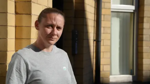 BBC Elissa Hubbard wearing a grey Nike T-shirt and looking at the camera. She is standing in front of her home which is yellow brick and a window is visible in the background