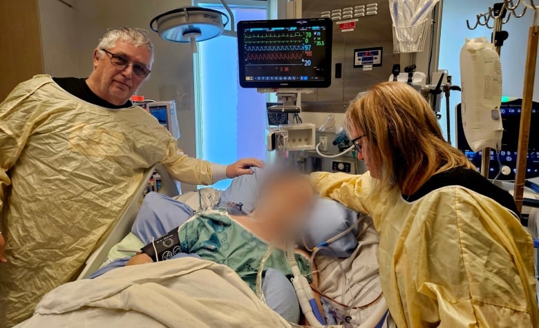 An older man and woman stand next to a hospital bed where a figure is surrounded by tubes and equipment. The patient's face is blurred to protect their privacy. 