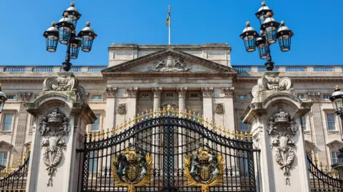 Getty Images Exterior of Buckingham Palace. It is a large stone-coloured building with columns and a balustrade at the top. In the foreground, there are two large pillars with lanterns on top of them and ornate black and gold gates in the middle. 