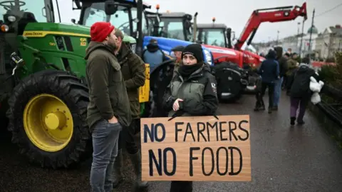 Getty Images A female dressed in a green and black rain coat holds a sign saying No Farmers No Food. A crowd of people and Green, red and blue tractors are in the background.