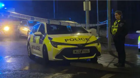 BBC A female police officer stands next to a police car next to police tape connected between two lampposts, with other emergency vehicles seen in the background. 