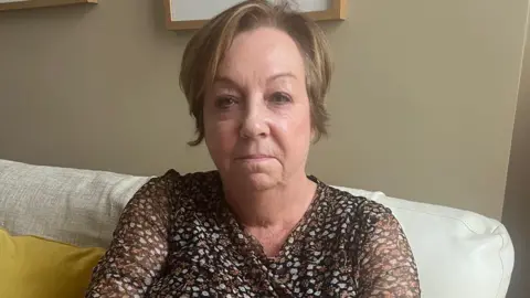 BBC Deborah Crisp, a woman with short light brown hair wearing a brown patterned top. She sits on a white sofa next to a yellow cushion, in front of a beige wall.