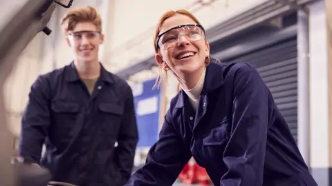 Getty Images Two young apprentices, one male and one female, smile with looking at a car engine while wearing blue jumpsuits and glasses