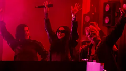 Getty Images Romy, Charli XCX and Robyn perform during day three of Glastonbury Festival 2024. The pop stars all have their hands up as they stand behind the DJ decks. The're lit by a red light and Charli is wearing her trademark sunglasses. 