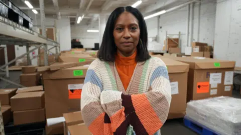 BBC Handbag designer Sherrill Mosee, wearing a bright orange turtleneck, stands with her arms crossed in a warehouse with boxes in the background