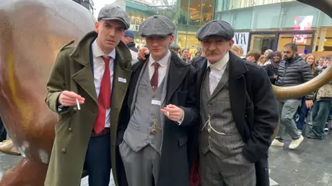 BBC Three men in grey flat caps and overcoats. two of them are holding cigarettes 