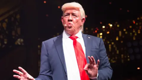 Mike Osman Mike Osman dressed as Donald Trump. He is wearing a blonde wig and pursing his lips together. He is wearing a blue suit and red tie.