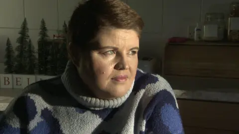 BBC Emma - a woman with short brown hair has her head turned from the camera looking out the window as sun lights up her face. She is sitting down in a kitchen and is wearing a grey turtleneck jumper with large blue spots.