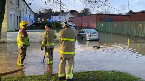 PA Media Three firefighters in uniform on residential streets in Belfast conduct a search as a cars tyres are submerged