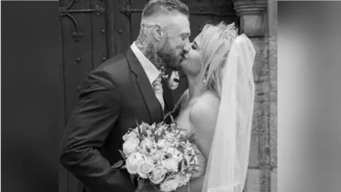 Family photo Aaron and Samantha Morris on their wedding day. It is a black and white photo and they are kissing while smiling outside a church door. The bride has bare shoulders, a tiara, white long veil and is holding a bouquet of roses.