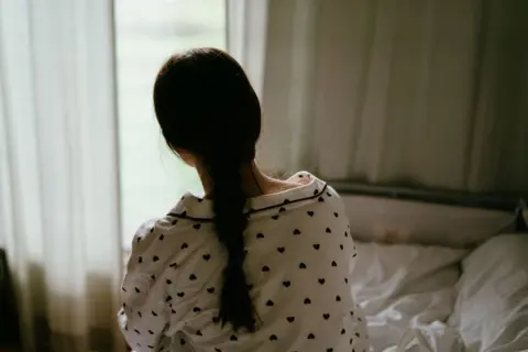 Getty Images A teenage girl wearing pyjamas sitting on a bed, with her back to the camera, looking out towards a window