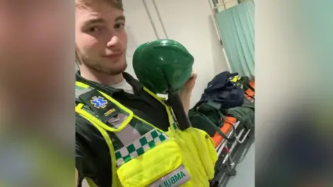 Facebook Jamie Kadolski poses in a selfie wearing an high-visibility vest with "ambulance" written on it. Underneath the vest is a green polo shirt and he is holding a green helmet. He is has a radio attached to the vest. In the background is a stretcher with orange mattress with bags on top. 