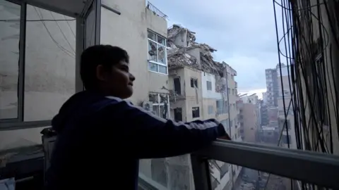 BBC Boy looks out from a balcony, facing right. Damaged buildings can be seen.