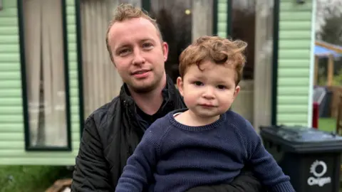 BBC Eoin McGovern is standing outside a green mobile home and is holding his young son