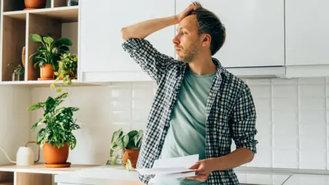 Getty Images Man standing in a kitchen with a bill in his hand and the other hand on his head.