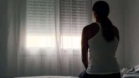 Getty Images Woman sitting on a bed staring out of a window covered by a net curtain