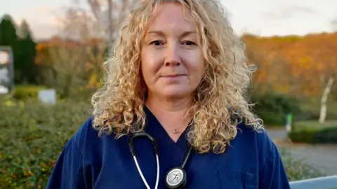 BBC Dr Samantha Jenkins wearing blue scrubs and with curly blonde hair looks at the camera