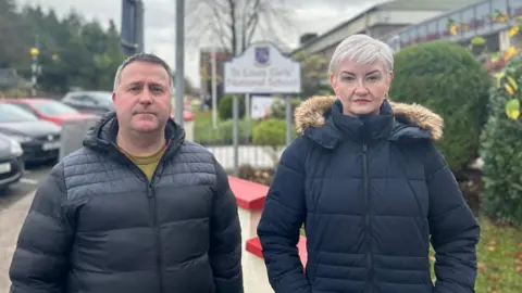 BBC Sean and Julieanne Lynch stand in front of a low level cream wall which has it's top painted bright red. Behind them is an out of focus sign for St Louis Girl's National School, where their daughter was last seen alive. Julieanne is wearing a black duffle coat with her hands in her pockets. She has short blonde hair and red lipstick. Sean, who has short dark hair, is stood to her left in a black padded coat. Both are looking at the camera. 